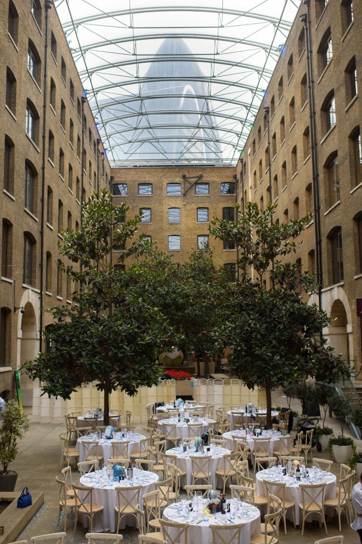 A wide shot of Devonshire Terrace, showing it's high glass ceiling above a courtyard containing two large trees, giving the venue an indoor/outdoor feel. Through the glass ceiling you can see the shape of the Gherkin, an iconic London landmark.
