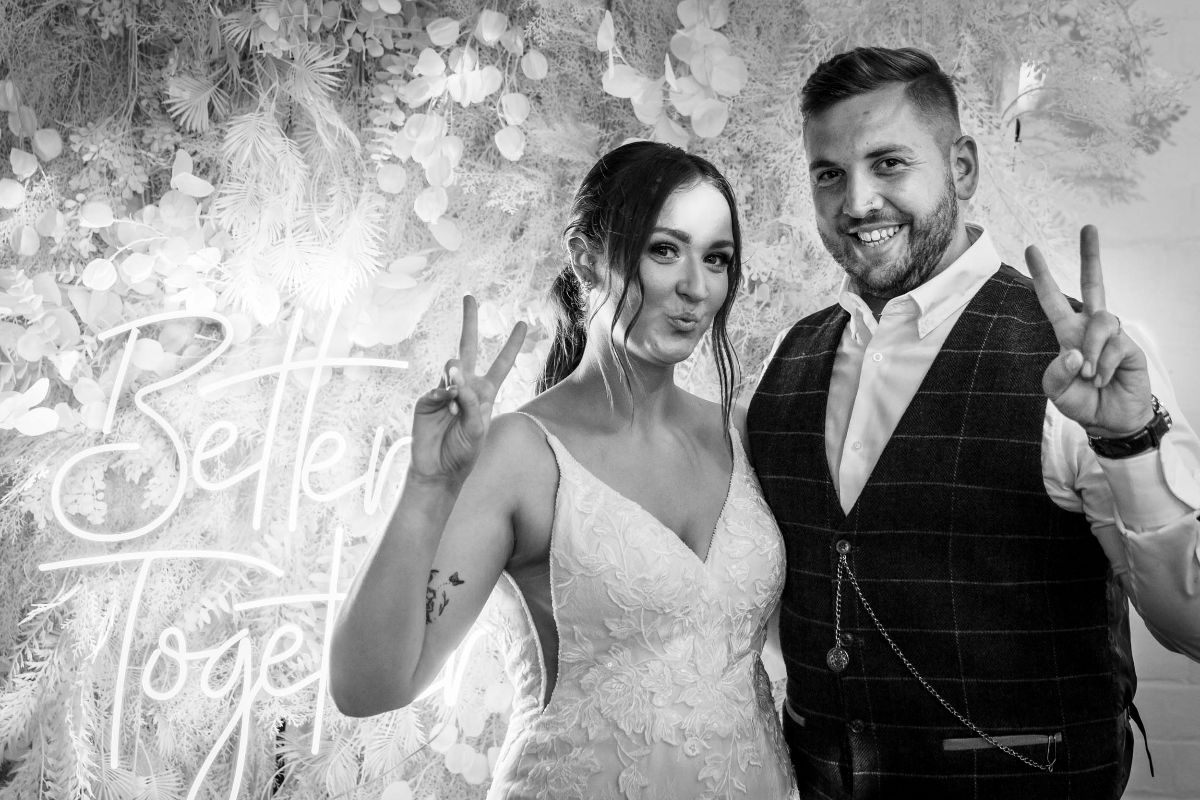 The Bride & Groom pose for a photograph in front of their lit flower wall at The Factory Hinckley